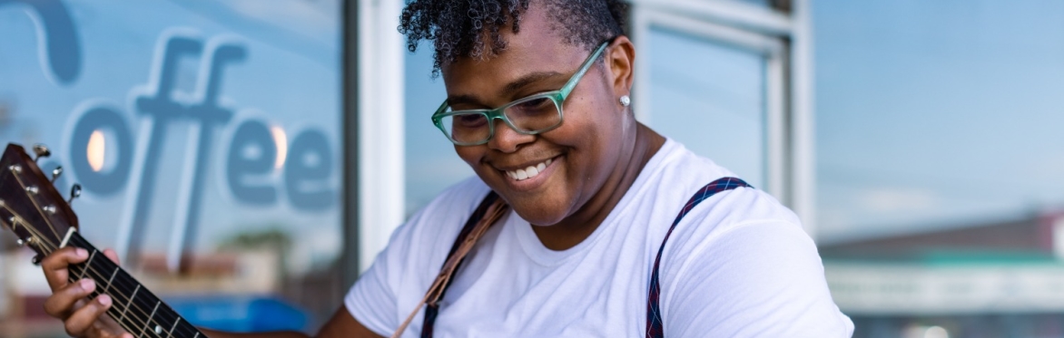 Crys Matthews smiles while playing an acoustic guitar outdoors, wearing glasses and a white shirt, ahead of her May 2 concert at the Sherbino Theater