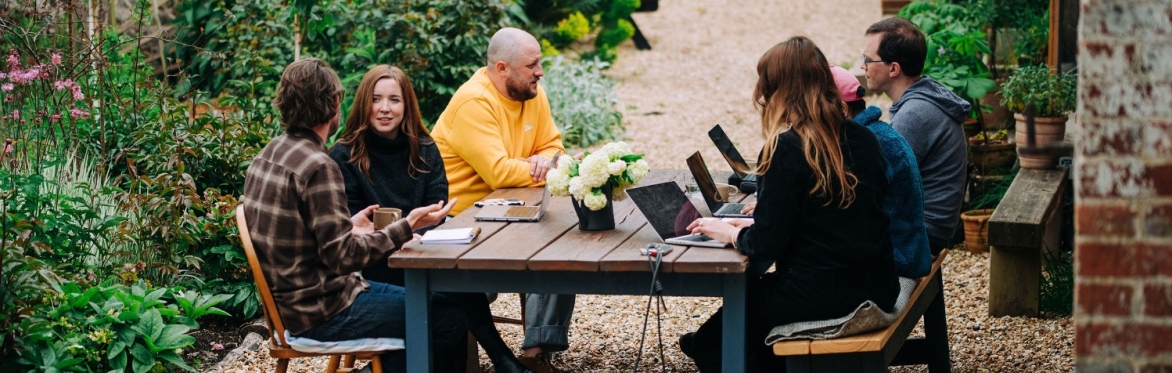 6 people working on laptops, drinking tea, deep in conversation. Sat around a wooden table, outside a flint barn, surrounded by flowers and nature.