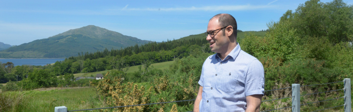 Rodge, a man in a short sleeved blue button down shirt, poses with his hands in his pockets. He is looking away from the camera. It is a bright sunny day and there is a lush, green landscape.