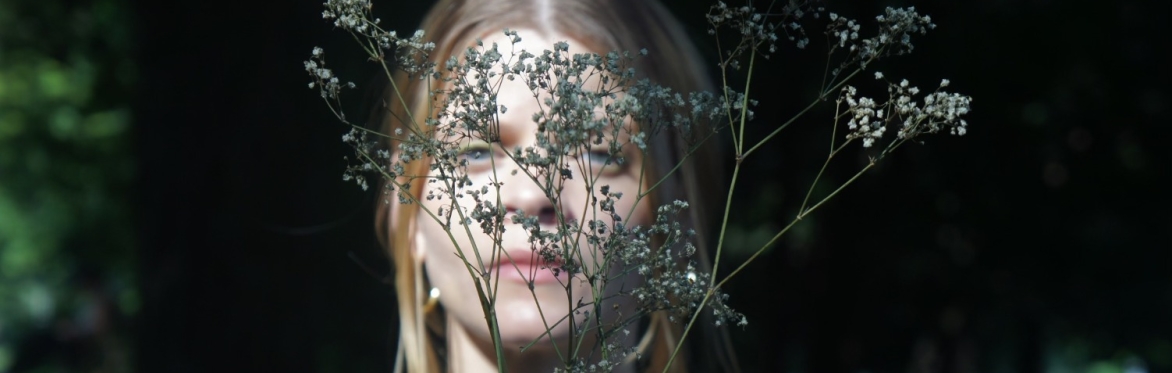 A person with long blonde hair, wearing a sleeveless black top, stands outdoors holding up a delicate cluster of dried wildflowers in front of their face. The flowers obscure much of their facial feat