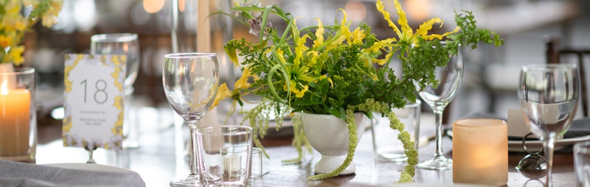 Image id: A detail image of a beautifully set table at the 2024 New City Arts Spring Dinner table. In the foreground, a wooden table features a floral centerpiece in a small white vase with vibrant gr