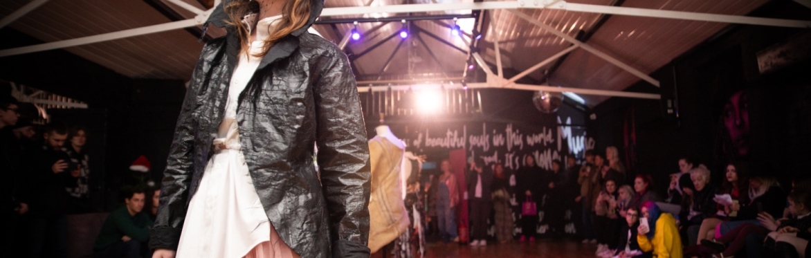 female model on the catwalk wearing black leather jacket, black mesh over her face, a white top and a pink tulle skirt