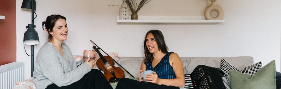 Rachel Campbell and Holli Scott are sitting with their feet up facing each other on a sofa. There is a fiddle next to Rachel and an accordion next to Holli. They are both holding a mug and laughing.