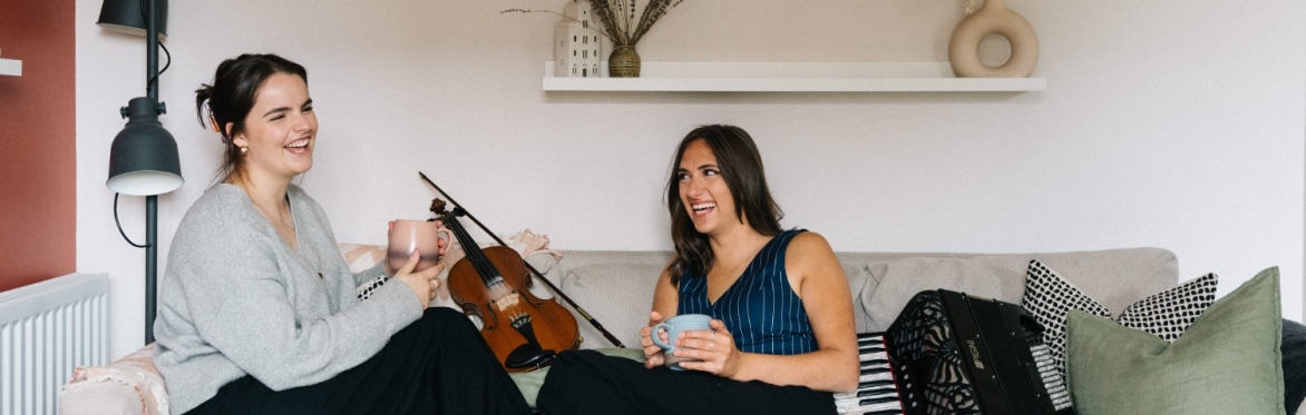 Rachel Campbell and Holli Scott are sitting with their feet up facing each other on a sofa. There is a fiddle next to Rachel and an accordion next to Holli. They are both holding a mug and laughing.