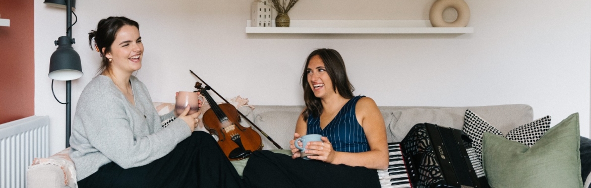 Rachel Campbell and Holli Scott are sitting with their feet up facing each other on a sofa. There is a fiddle next to Rachel and an accordion next to Holli. They are both holding a mug and laughing.