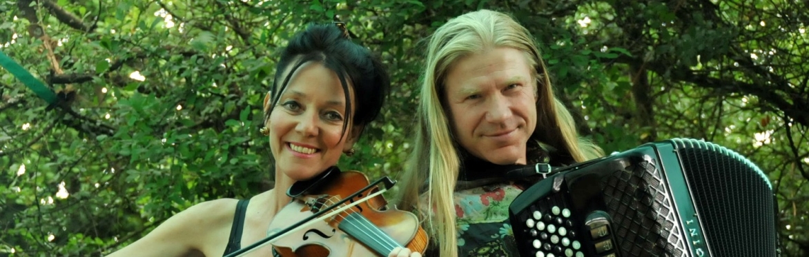 Two musicians outdoors, a woman playing violin and a man playing accordion, smiling at the camera.