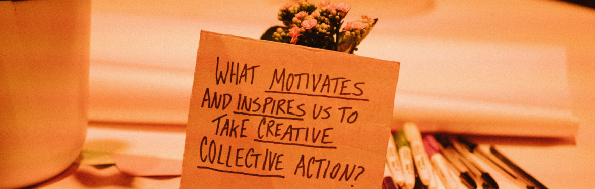 A cardboard sign reading 'What motivates and inspires us to take creative collective action?' with a plant and pens in the background.
