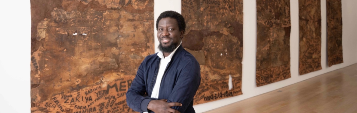 An image of the artist Ibrahim Mahama in front of his large tattoo paintings
