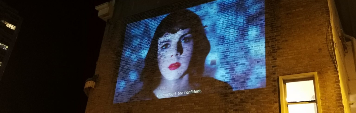 A projection onto a large wall showing a white trans woman with a black bob hairstyle and wearing red lipstick.