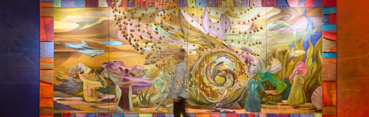 A photograph of someone walking through a colourful art installation, with a large plinth and hanging gold balls.