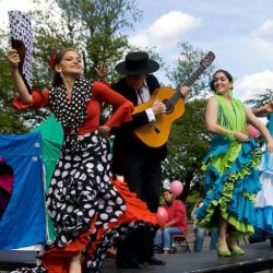 Performance action shot, set on an open-air stage: a flamenco dancer with a spotty dress and spotty fan is accompanied by a flamenco guitarist in a dark suit and a Spanish  wide-brimmed hat.
