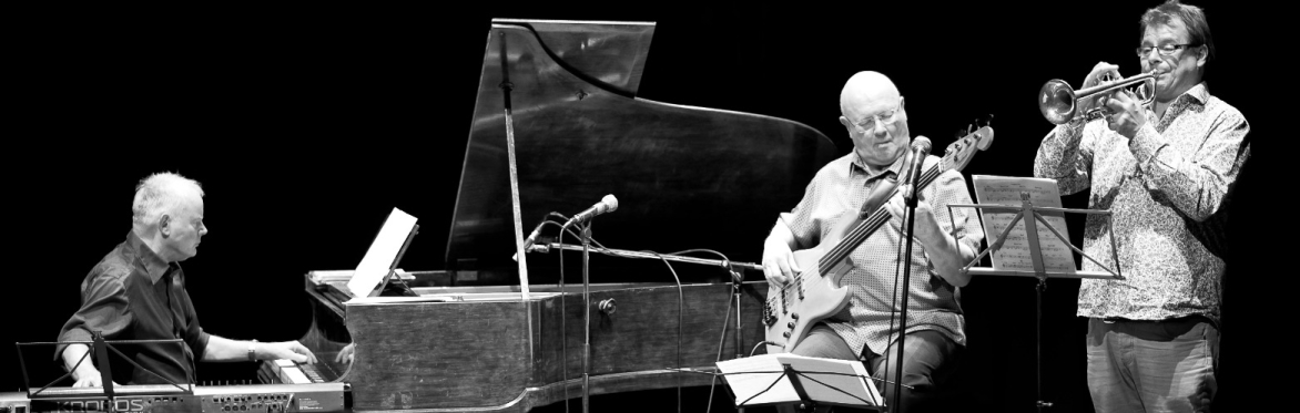 Black-and-white photo of a jazz trio performing on stage. A pianist, bassist and trumpeter play beneath spotlights, with music stands, microphones and audience silhouettes in the foreground.