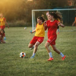 Four girls play rugby on a grassy field at sunset. One carries the ball while others move to tackle her, showing teamwork and determination.