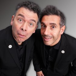 Two men in black suits lean toward the camera with playful, surprised expressions.