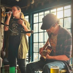 Fox Cub and The Big Red perform an intimate live acoustic set inside a warmly lit pub. One singer stands at a microphone while a seated guitarist plays beside a sunlit window, with drinks and mic