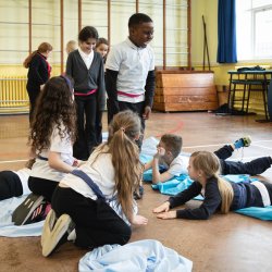 Children using blue fabric to represent a river during a creative storytelling and drama workshop in a school hall.