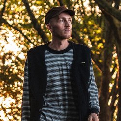 Dean Frost stands outdoors among tall trees at golden hour, wearing a dark cap, striped long-sleeve top and black gilet. He looks thoughtfully into the distance as warm sunlight filters through the wo