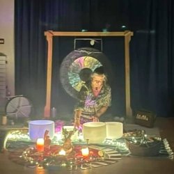 Faye sitting in front of a gong, playing crystal singing bowls