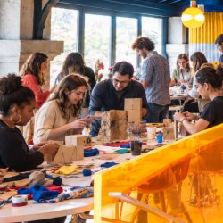 Group of people being creative around a table filled with paint and cardboard, while others in the background are having a glass of wine during a Creative Playground Night.
