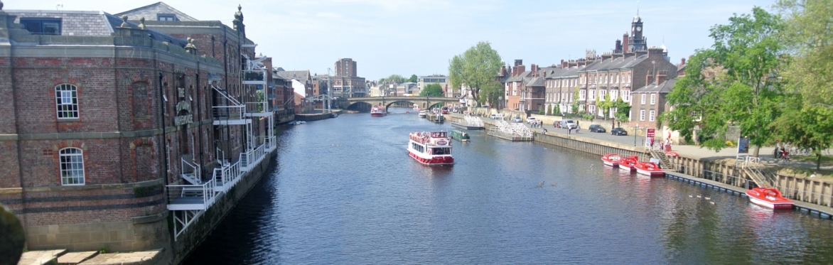 View along the River Ouse in York on a bright day, with a sightseeing boat on the water, historic riverside buildings, trees and boats moored along the bank. The photograph is taken from Skeldergate B