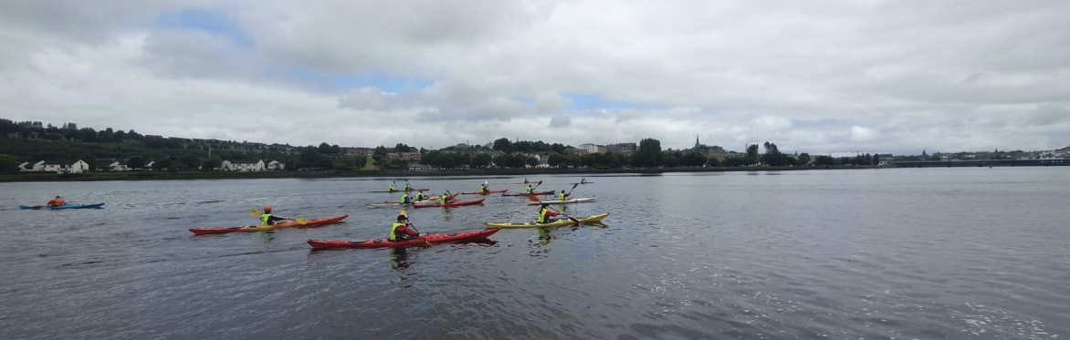 A group of kayakers wearing high-visibility vests paddle colourful kayaks across a wide river under a cloudy sky, with a town and shoreline visible in the distance.