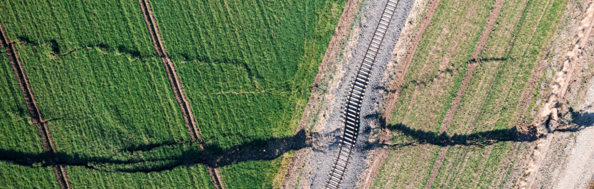 Aerial view of green fields with a dark crack running through them, crossing a railroad track. The image conveys tension and disruption.