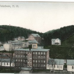 Colorized vintage postcard showing a complex of brick and clapboard mill buildings in a green, hilly landscape.