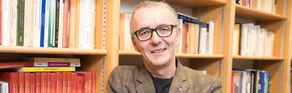 A man with short grey hair wearing glasses, stands with his arms folded, in front of a bookcase filled with textbooks.