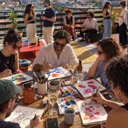 Group of people drawing and painting around a table filled with paint and paper, while others in the background are having a glass of wine with a view over Lisbon, during a Creative Playground Night.