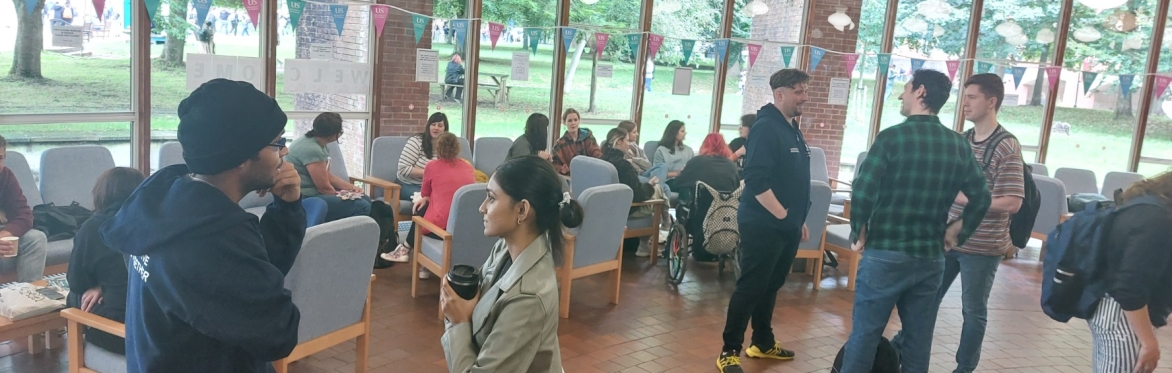 A photograph of a room of people. Some are sitting on comfortable chairs, others are standing, they are talking and smiling and look relaxed.