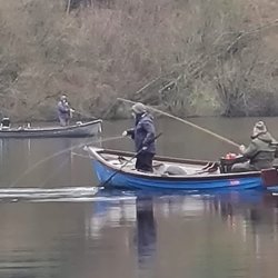 Two people in a blue and white rowboat on a calm lake, with one person standing and using a long pole. A second boat and a wooded shoreline are visible in the background.