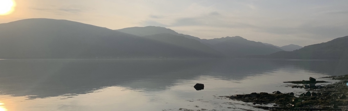 A hazy landscape of low sun over rolling hills and their mirrored reflection in a lake