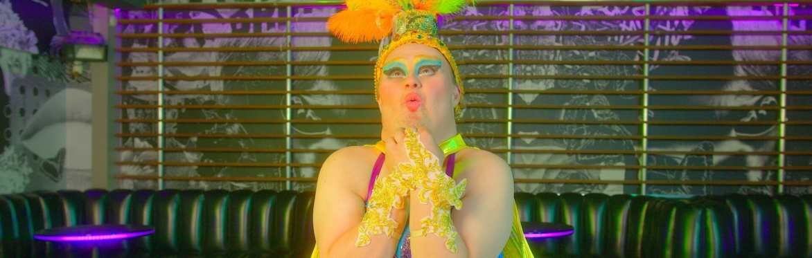Performer in colourful drag costume poses in a nightclub, wearing a sparkling headdress with bright rainbow feathers, dramatic blue eye make-up, gold gloves and a vivid green and blue outfit.