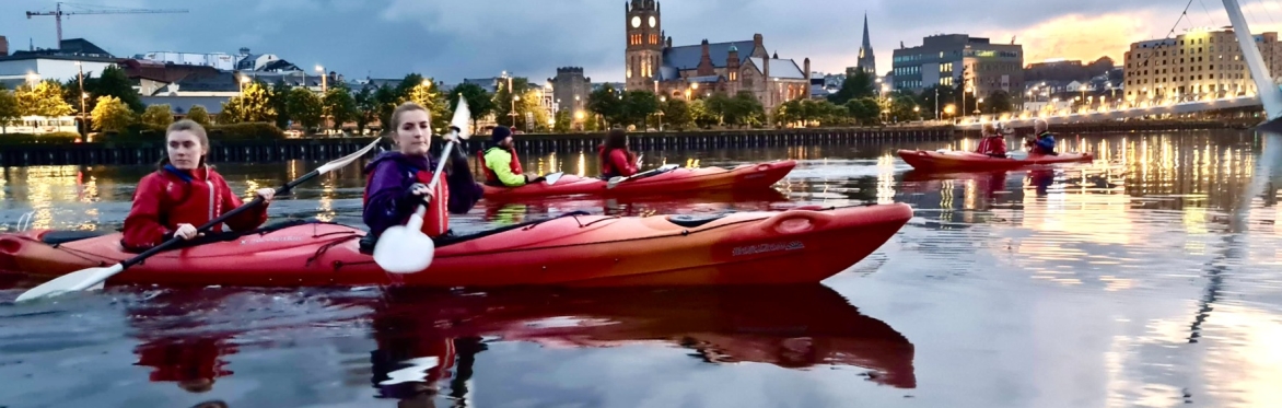 Kayakers on a river at dusk with city lights.