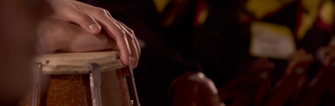 A close-up film still of a hand resting on the skin of a small drum, its fingers curved gently over the edge mid-play. The warm, amber tones of the drum's body contrast with the dark, softly blurred b