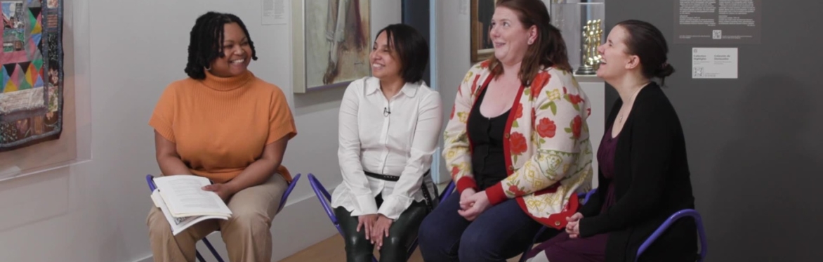 Four women sit on stools in a gallery of the Smithsonian National Portrait Gallery. Three of the women are facing towards the top right corner of the photo, considering a quilt