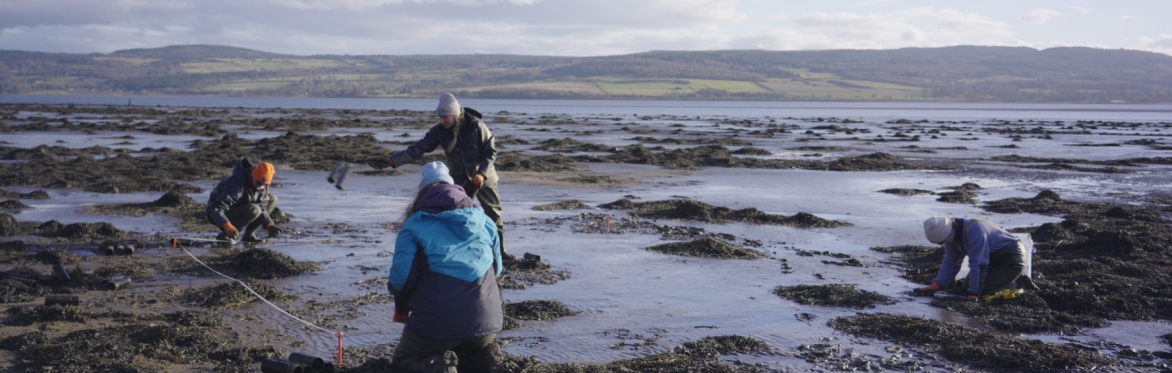 6 smiling people standing on a beach in waterproofs and hats. The one in the middle is holding a core of sediment and seagrass.