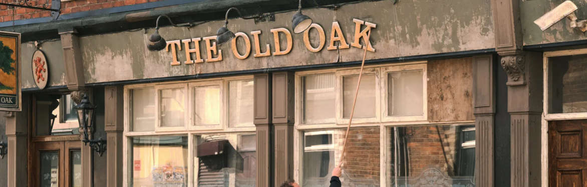 A still from the film featuring an old pub front, with a sign that reads 'THE OLD OAK'. The final letter K is falling, and a person is trying to fix it with a long stick