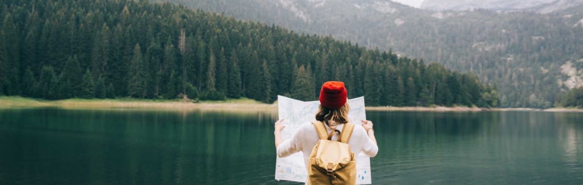 Person with their back to us looking at a map. They are facing a lake with trees and hills behind it.