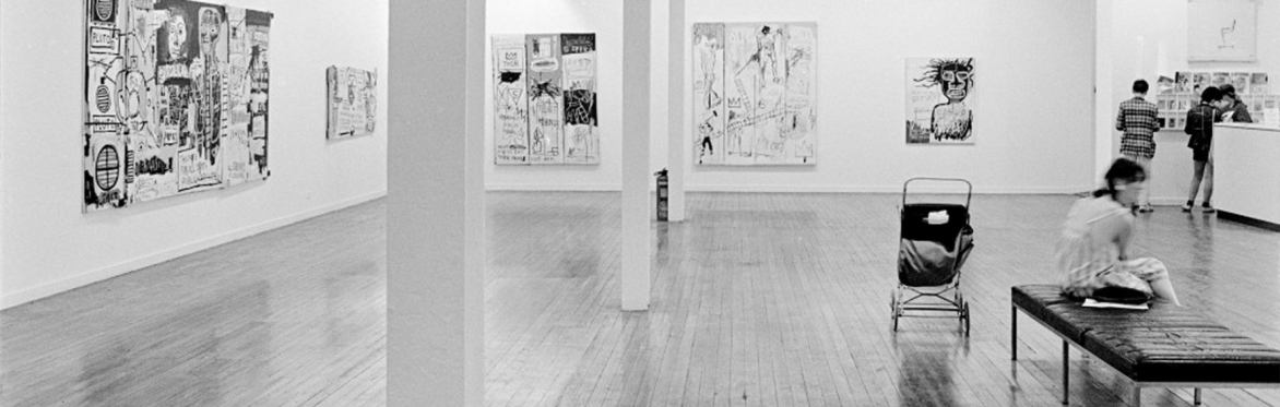 A black and white photo of an exhibition space and a child's buggy in the Frutimarket in 1984. There is a buggy in the space and people milling around the Basquait exhibition
