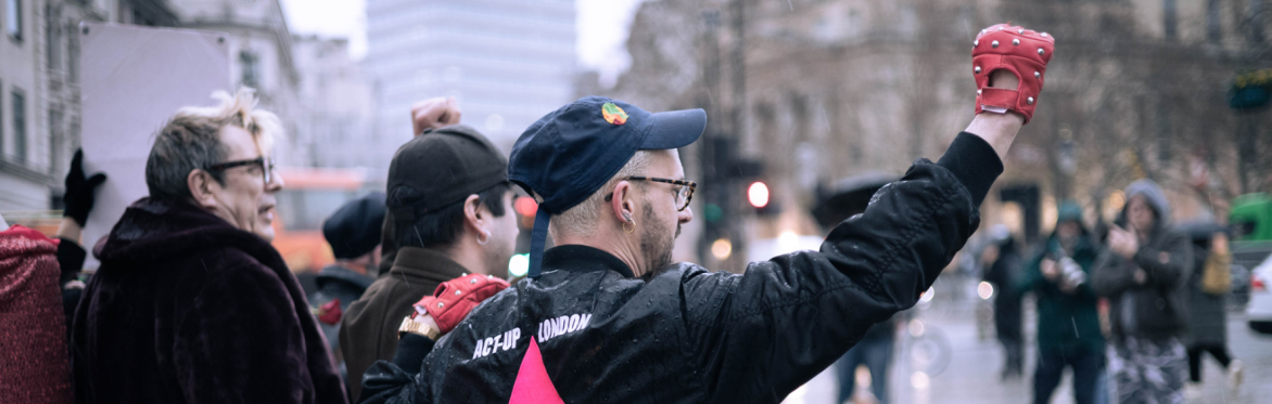 A group of people protest outdoors in a city; one person raises a red gloved fist, wearing a jacket with a pink triangle and the words "ACT-UP LONDON"