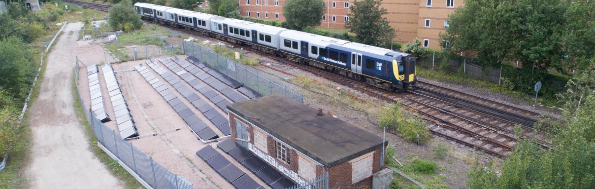 In the foreground, there is a small solar installation next to a single storey building, and in the background a train is going past along the railways.