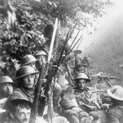 Black-and-white photograph of First World War soldiers wearing steel helmets, gathered in a trench with rifles and bayonets.