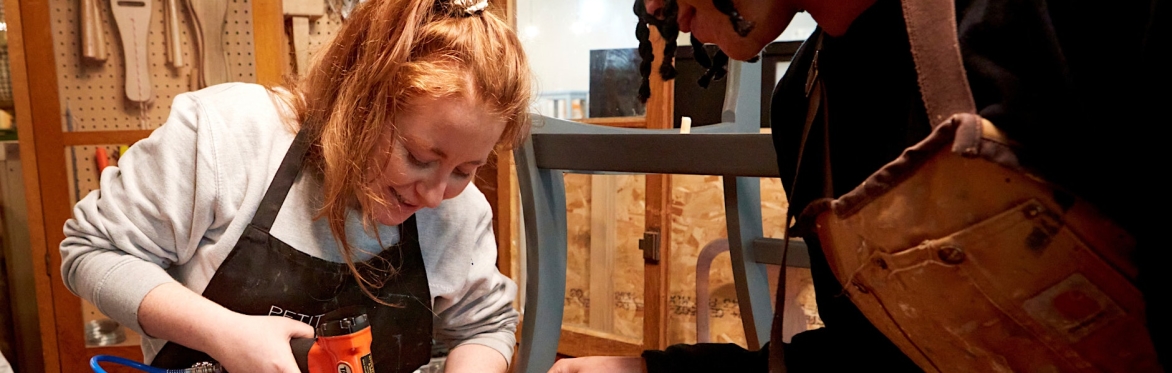 2 people are in a workshop setting, the one on the right is directing the other person how to upholster a dining chair seat with black fabric, using an air powered staple gun.