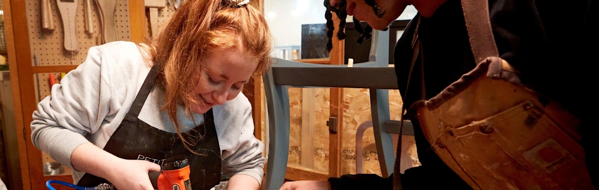 2 people are in a workshop setting, the one on the right is directing the other person how to upholster a dining chair seat with black fabric, using an air powered staple gun.