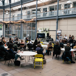 A gathering of people clustered around small tables listening to speakers on the stage to the right of the image