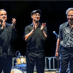 **Alt text (under 200 characters):** Three musicians stand on stage acknowledging applause. Two clap while the third smiles, with drums and stage equipment behind them under concert lighting.