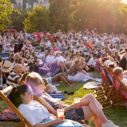 A crowd of people sit in deckchairs in a park in the evening sunshine.