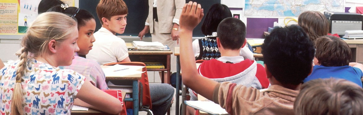 A classroom of children look at a boy with his hand up.