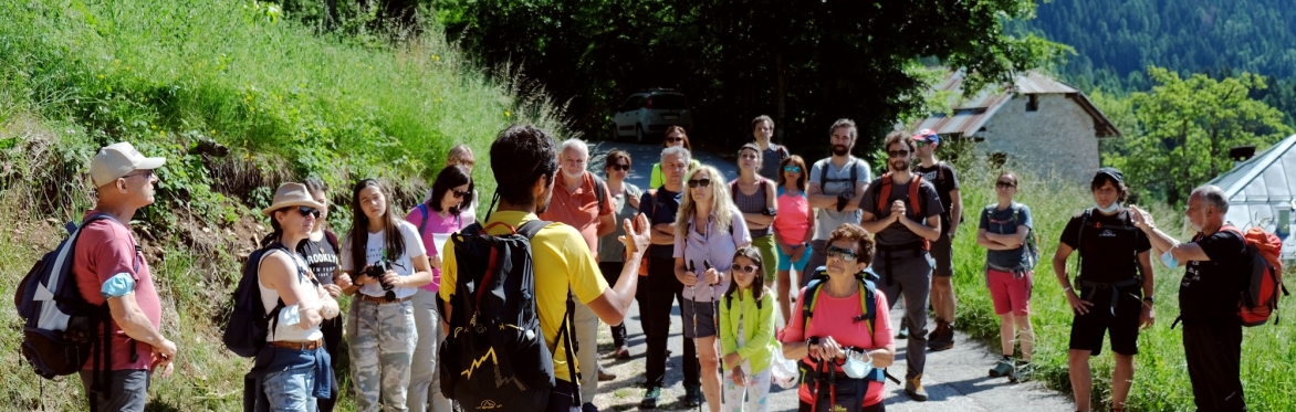 Gruppo di persone ferme ad ascoltare Luca Stefenelli. Sono su una strada asfaltata all'inizio di un sentiero di montagna. Poco lontano si intravedono due edifici (di cui una casa e una serra)